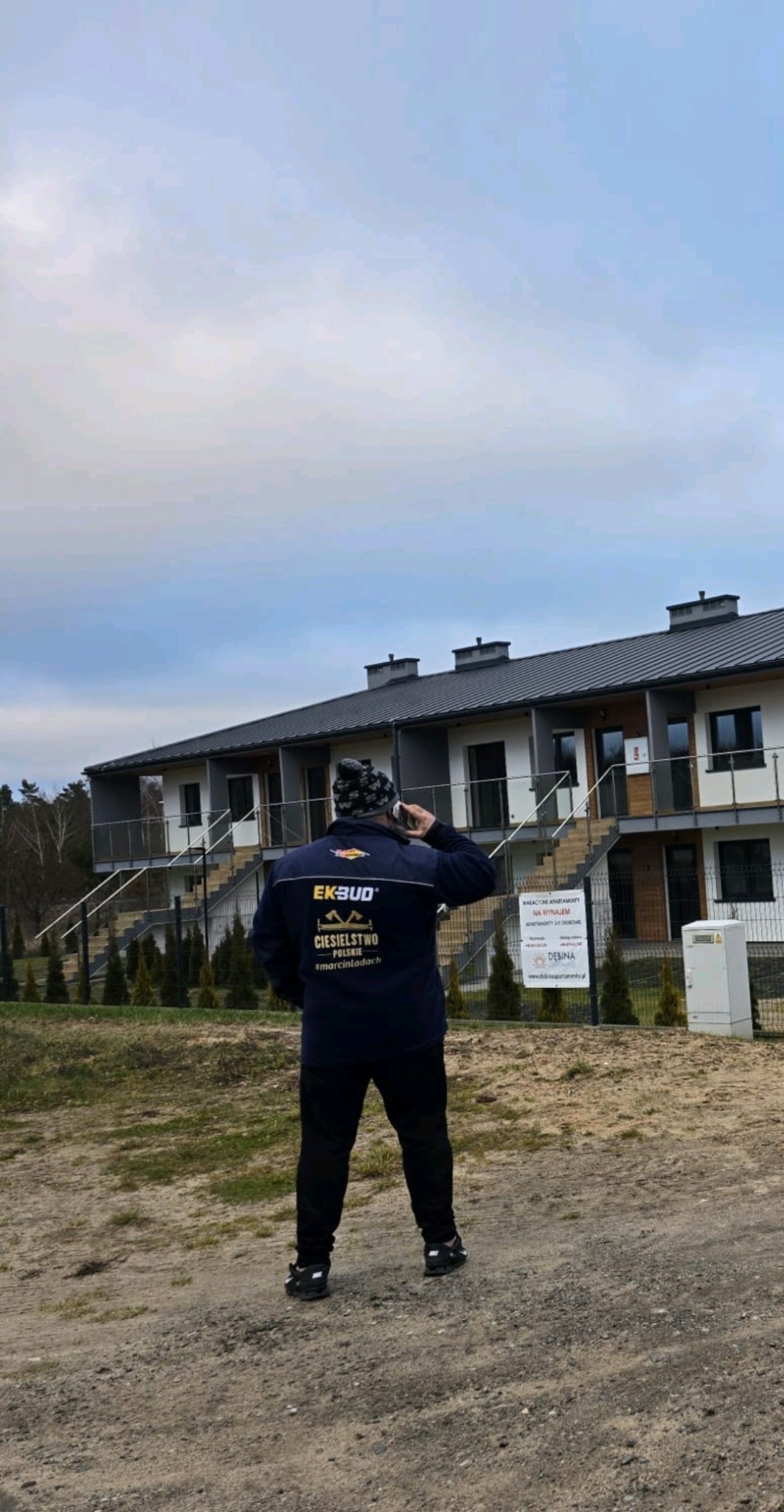 Mężczyzna w kurtce z logo firmy dekarskiej EKSBUD rozmawia przez telefon na tle szeregu nowo wybudowanych domów z ciemnymi dachami i obróbkami blacharskimi.