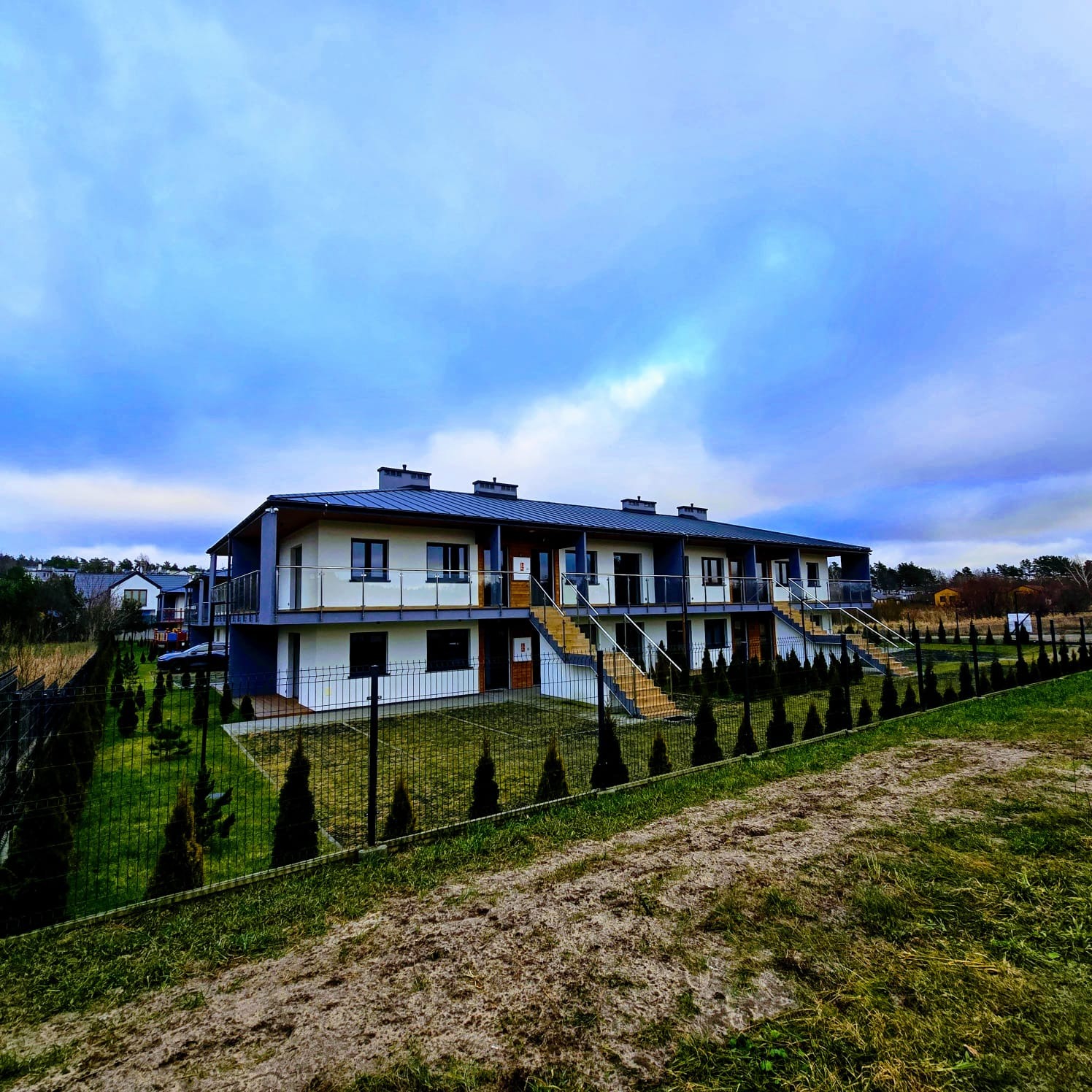 Dwukondygnacyjny budynek wielorodzinny z szarym dachem i balkonami, widoczny zza metalowego ogrodzenia i rzędu małych drzewek iglastych.