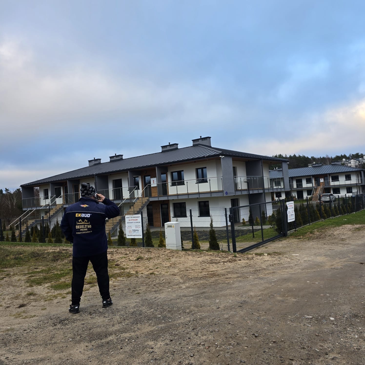 Mężczyzna w kurtce firmowej Ek-Bud stoi tyłem na żwirowej drodze, rozmawiając przez telefon, na tle nowoczesnych, dwukondygnacyjnych domów szeregowych z szarymi dachami i białymi elewacjami...