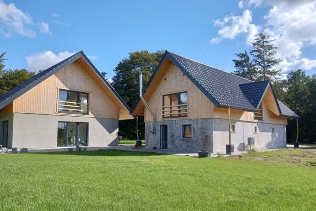 Dwa identyczne, parterowe domy w stanie surowym zamkniętym, z ciemną dachówką i drewnianą elewacją na poddaszu, na tle zielonego trawnika i drzew.