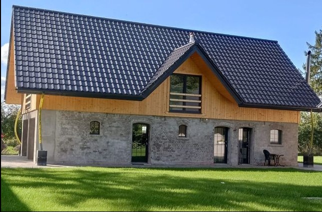 Nowoczesny dom z ciemną dachówką, drewnianą elewacją i szarym murem, otoczony zielonym trawnikiem.