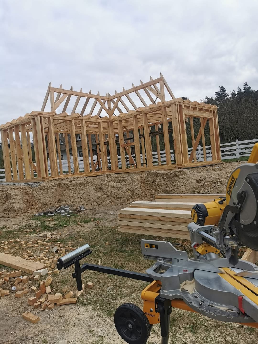 Konstrukcja drewniana domu w trakcie budowy, widoczna piła do drewna DeWalt, przygotowane deski i porozrzucane kawałki drewna na ziemi.