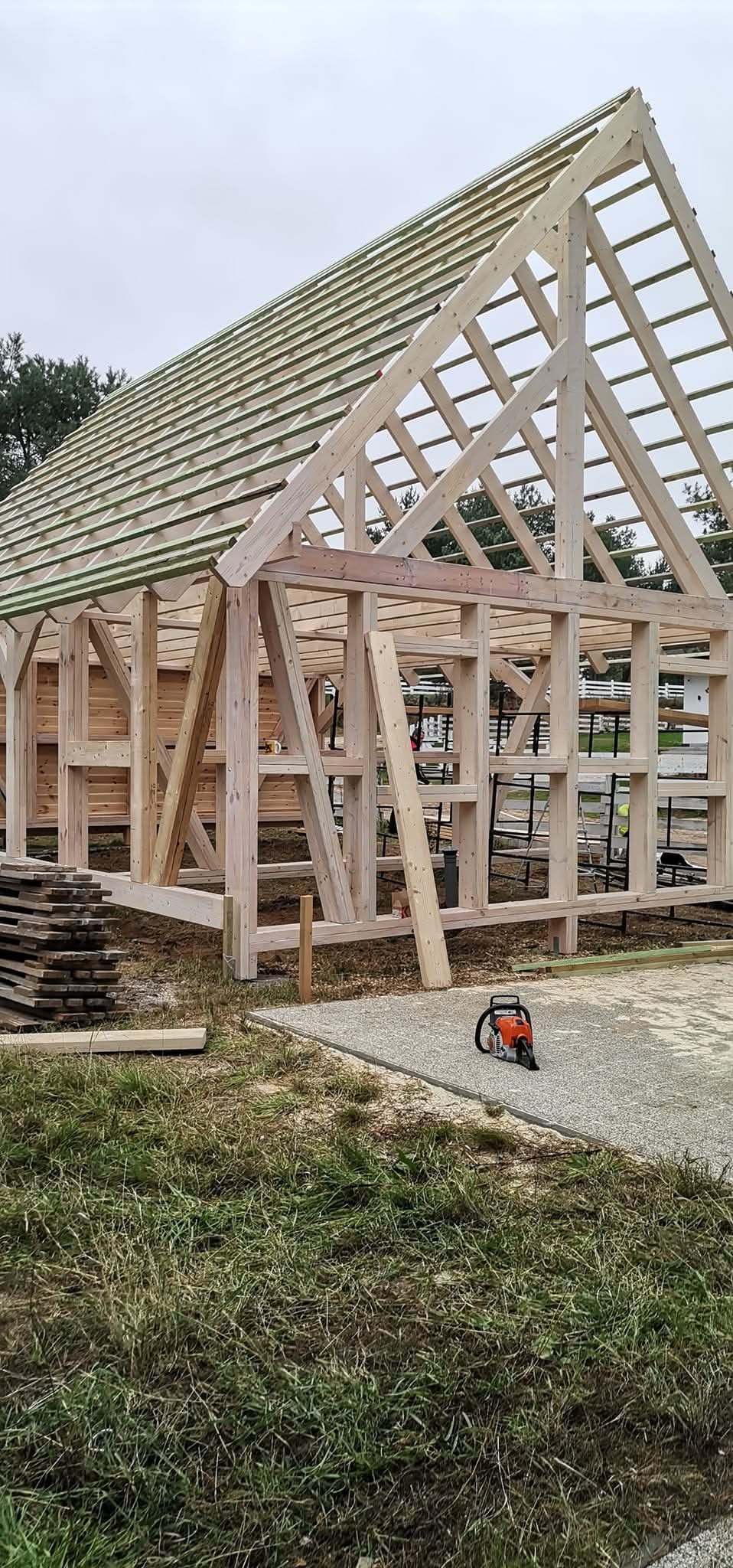 Konstrukcja szkieletowa domu w trakcie budowy, widoczne elementy drewniane, więźba dachowa i piła łańcuchowa na ziemi.