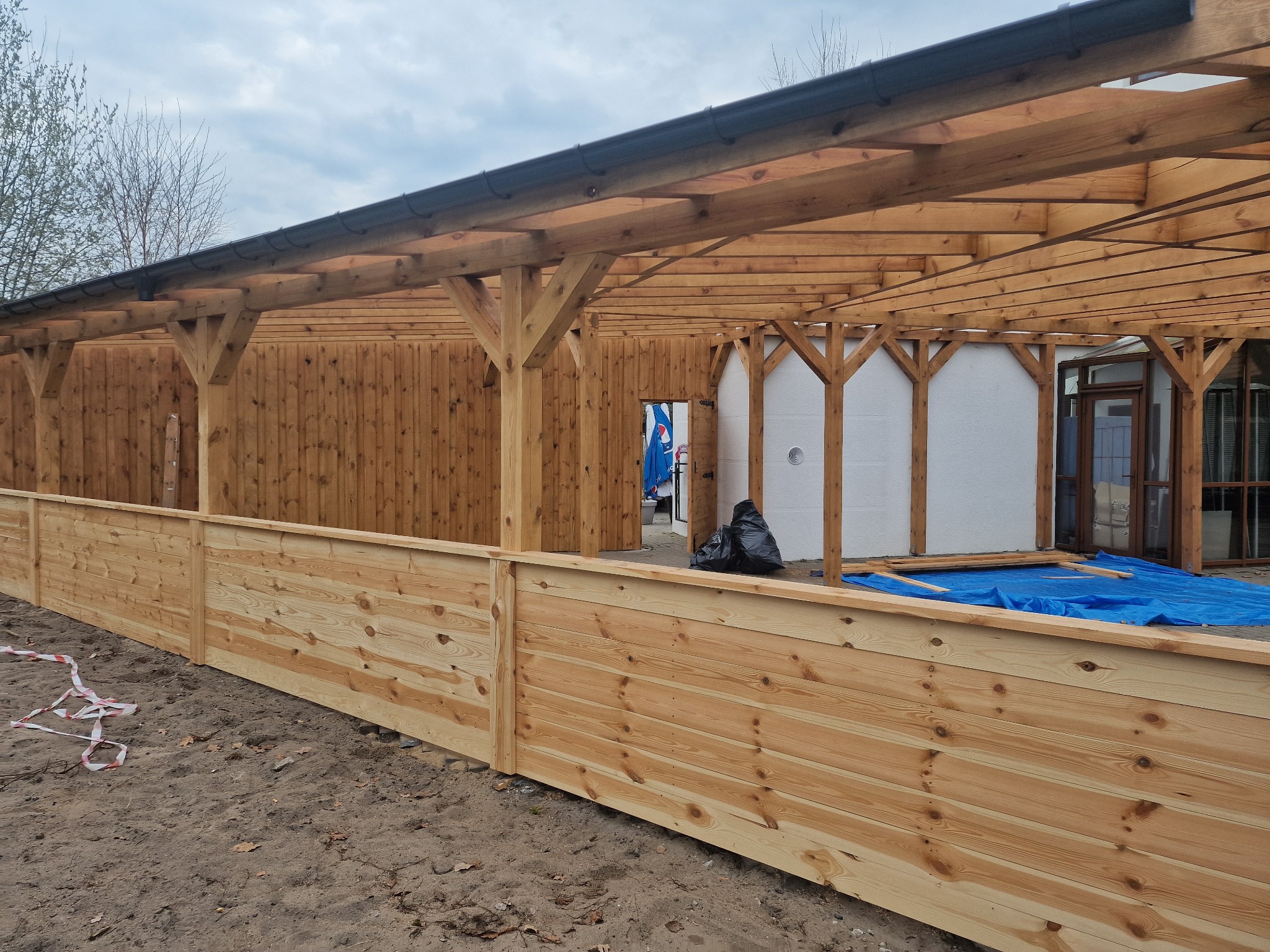 Drewniana konstrukcja zadaszenia tarasu w trakcie budowy, widoczne elementy więźby i obróbki blacharskie, teren budowy z taśmą ostrzegawczą.