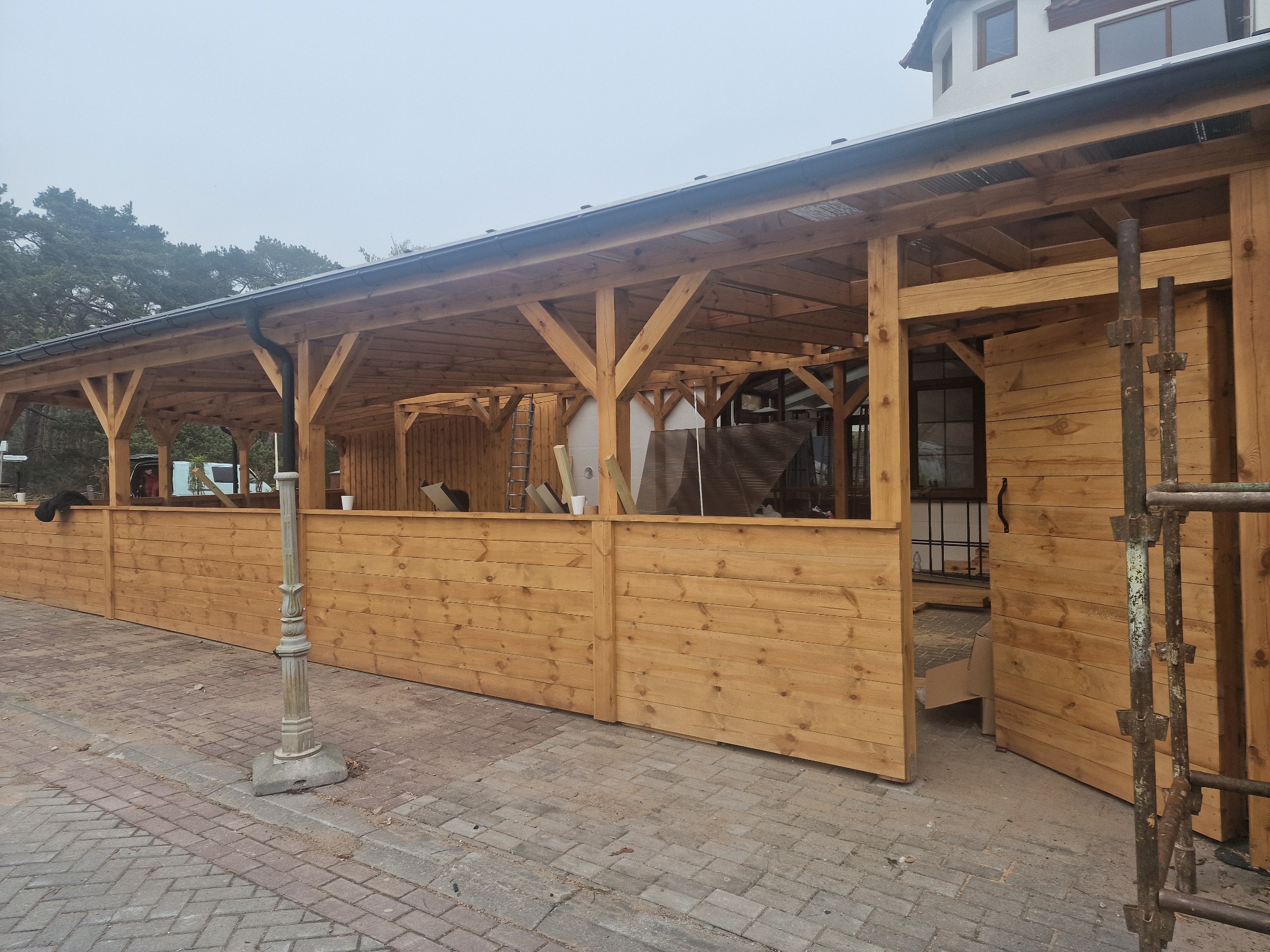 Drewniana konstrukcja z zadaszeniem, widoczne elementy więźby, ściany z desek, lampa uliczna, kostka brukowa. Ujęcie z zewnątrz, w tle zieleń i budynek.