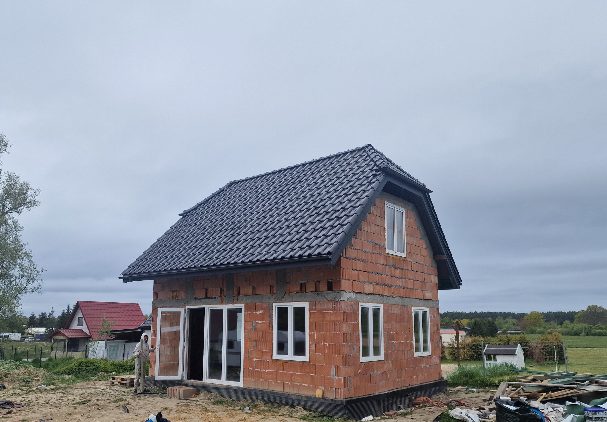 Niedokończony dom z czerwonej cegły z ciemnoszarym dachem z dachówki, białe okna, pracownik w drzwiach. Widok na okolicę i pochmurne niebo.