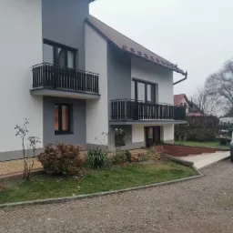 Dwukondygnacyjny dom z balkonami o szarej i białej elewacji, widok z zewnątrz, z podjazdem wyłożonym żwirem.