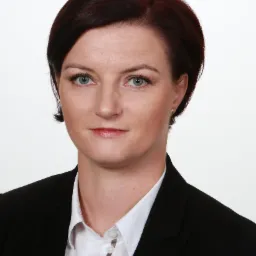 Portret kobiety w czarnym żakiecie i białej koszuli z subtelnym makijażem i krótkimi, ciemnymi włosami na białym tle.