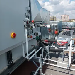 Kompleksowa instalacja wentylacyjna na dachu budynku z widocznymi przewodami, zaworami, manometrami i panelem sterowania z lampkami sygnalizacyjnymi, na tle błękitnego nieba z chmurami i zabudowy...