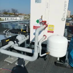 Instalacja przemysłowa Clivet na dachu budynku, widoczne białe urządzenie, rury, zawory, manometr, zbiornik wyrównawczy oraz elementy wentylacji. W tle zabudowa miejska.