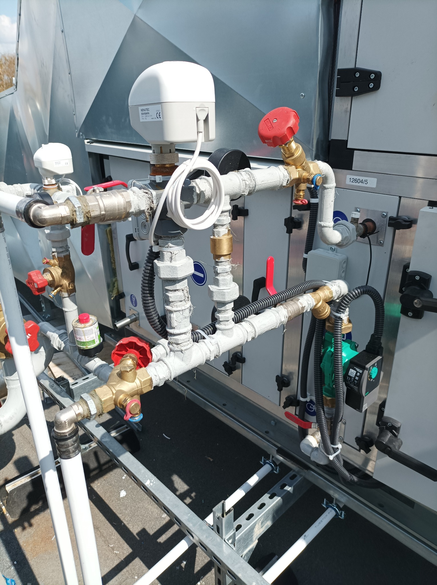 Złożona instalacja rur i zaworów na dachu, z elementami automatyki i sterowania, częściowo pomalowana na szaro, widoczne detale połączeń i etykiety.