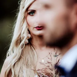 Portret panny młodej z intensywnym spojrzeniem, blond włosami i kwiatowymi kolczykami, częściowo zasłoniętej przez sylwetkę pana młodego, z widocznym tatuażem na dekolcie i delikatną biżuterią.