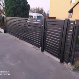 Nowoczesne, czarne ogrodzenie posesji z poziomych profili, brama wjazdowa, w tle budynek mieszkalny i samoch&oacute;d dostawczy.