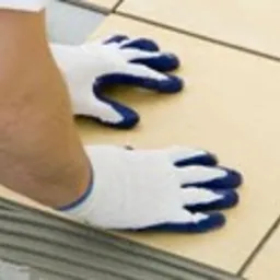 Układanie jasnych płytek podłogowych z użyciem zaprawy klejowej, widoczne ręce w białych rękawicach ochronnych dociskające płytkę do podłoża.