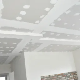 Wykończony sufit z płyt gipsowo-kartonowych z widocznymi łączeniami przed szpachlowaniem, z elementami dekoracyjnymi i oświetleniem w tle, w surowym stanie wykończenia wnętrza.