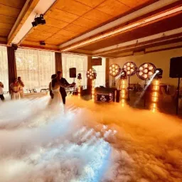 Para młoda tańczy pierwszy taniec w oparach dymu, z profesjonalnym oświetleniem i nagłośnieniem w tle. DJ za konsoletą. Dekoracja sali weselnej.