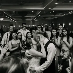 Czarno-białe zdjęcie pocałunku nowożeńców na weselu, otoczonych przez wiwatujących gości. W tle widoczne dekoracje sali i oświetlenie.