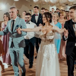 Przemysław Walkowski - Korowód weselny na parkiecie: para młoda w centrum, goście w eleganckich strojach, rustykalny klimat sali z dekoracjami i drewnianą podłogą, uśmiechnięte twarze.