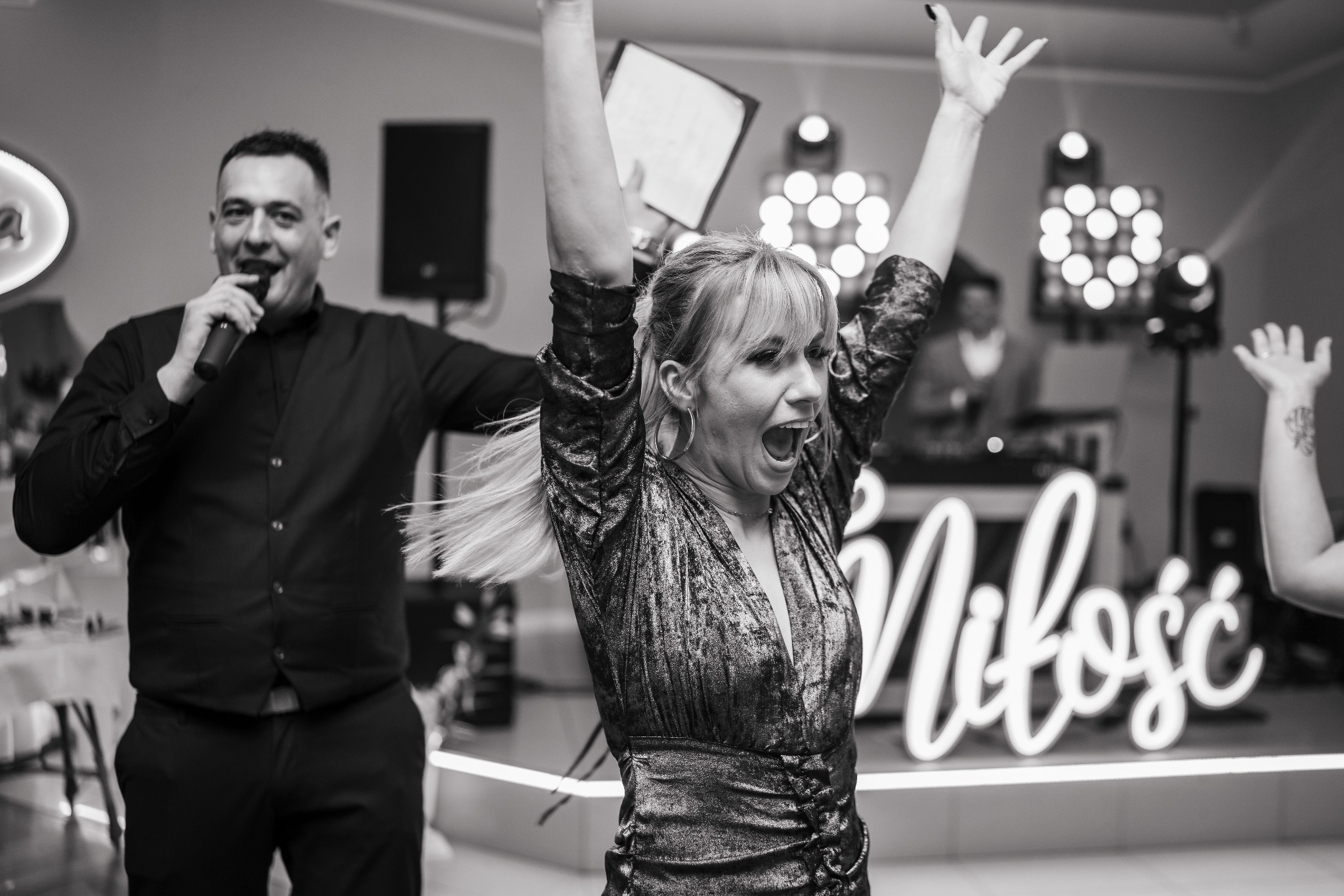 Czarno-białe zdjęcie z mikrofonem konferansjera i rozentuzjazmowanej kobiety na tle sceny z napisem 'Miłość' i oświetleniem, uchwycone w trakcie imprezy.