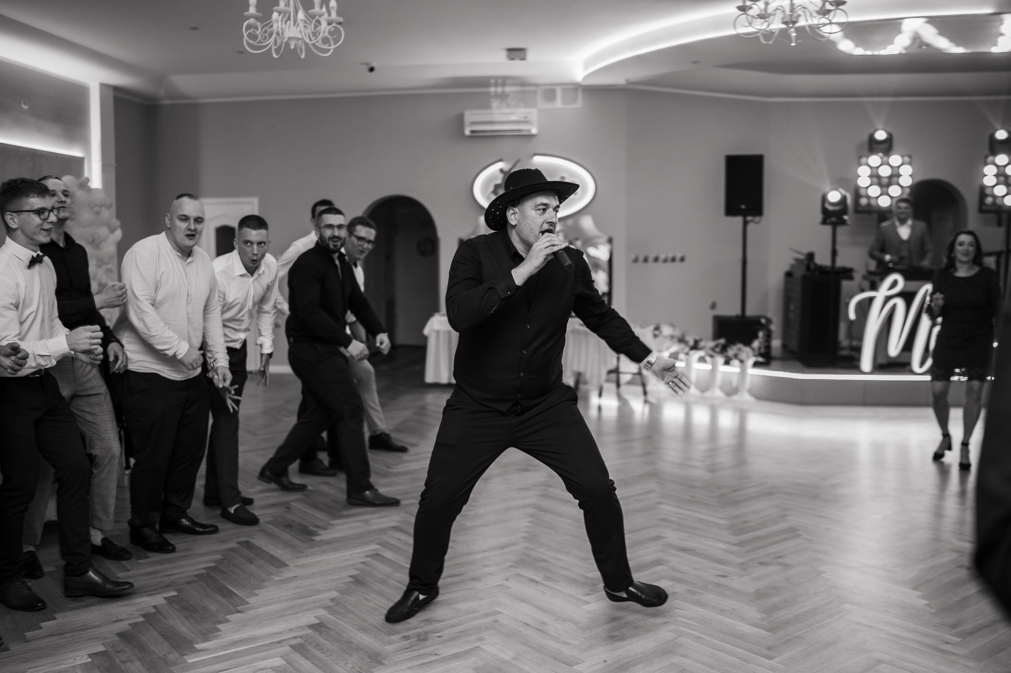 Wokalista w czarnym stroju, z mikrofonem, na parkiecie podczas wesela. Za nim zespół i goście, tworzący radosną atmosferę zabawy.