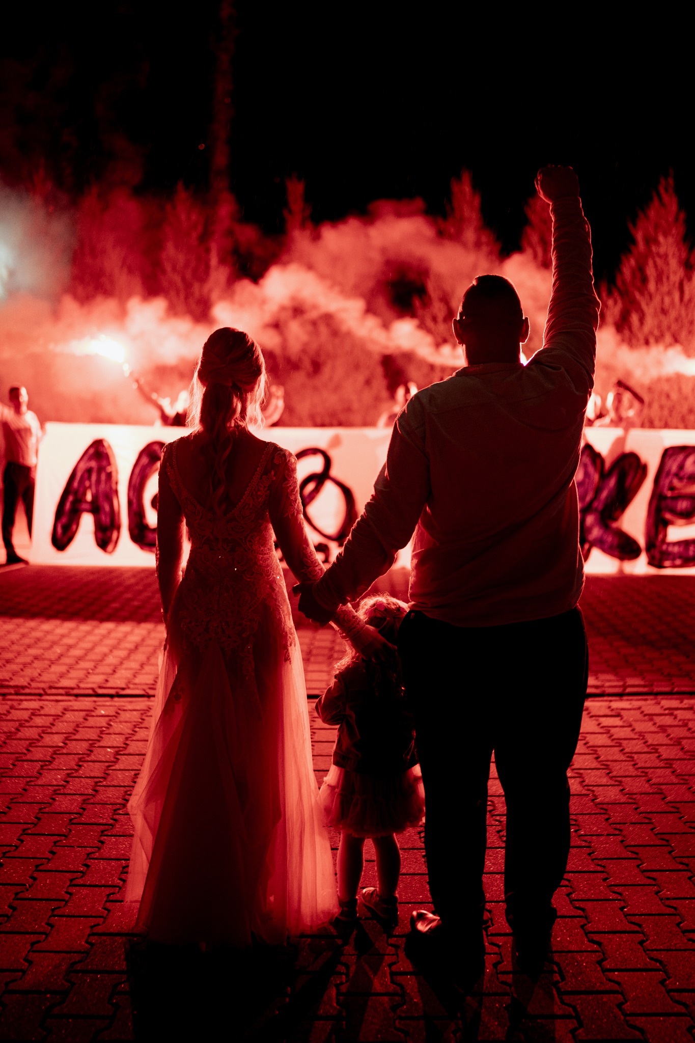 Para Młoda z dzieckiem, widziani od tyłu, trzymają się za ręce, a w tle czerwone race i baner z nazwą zespołu muzycznego, oświetlone intensywnym czerwonym światłem.