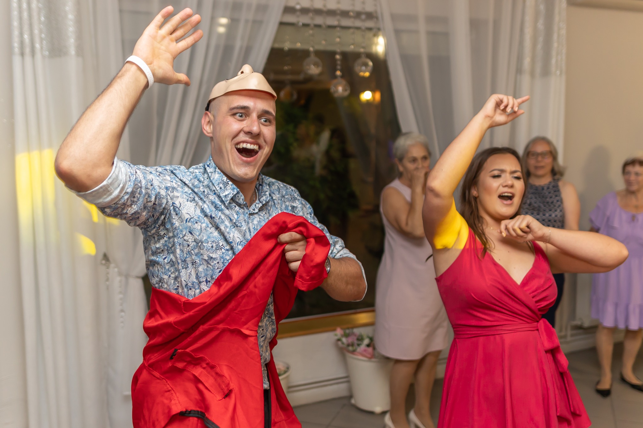 Wodzirej w masce z uniesioną ręką i kobieta w czerwonej sukience tańczą na imprezie. W tle okno i obserwujące osoby.