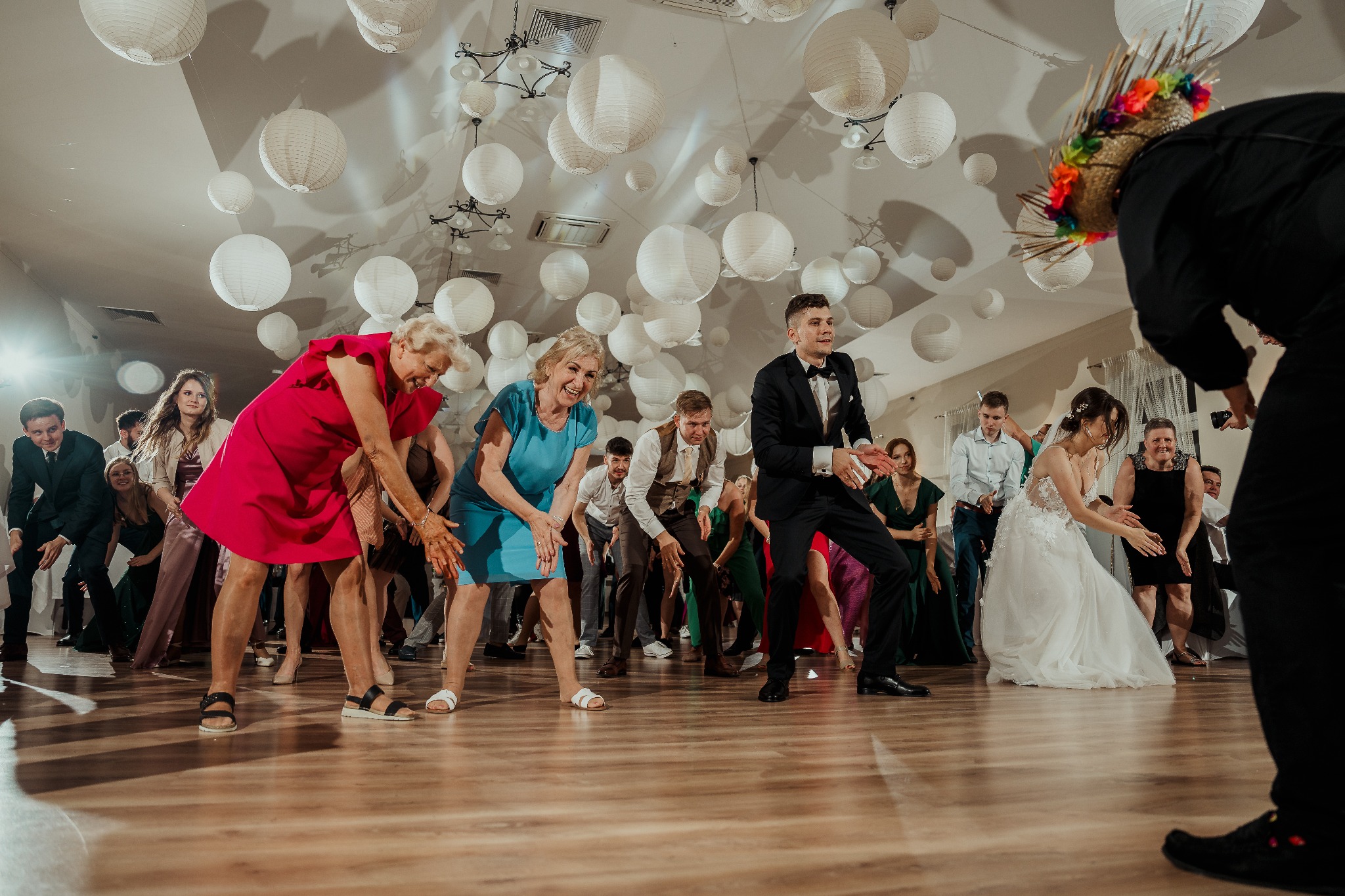 Rozbawieni goście weselni tańczą limbo pod laską trzymaną przez wodzireja w kolorowym kapeluszu. Dekoracje z papierowych lampionów pod sufitem.
