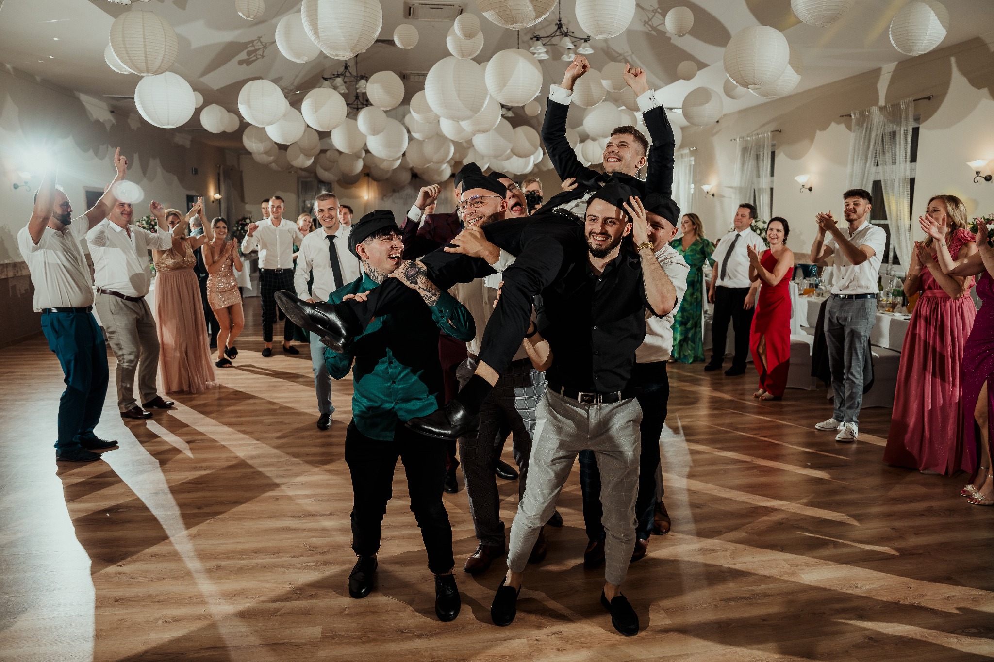 Rozbawiona grupa gości weselnych świętuje, unosząc w górę członków zespołu muzycznego na parkiecie, pod dekoracją z białych lampionów.