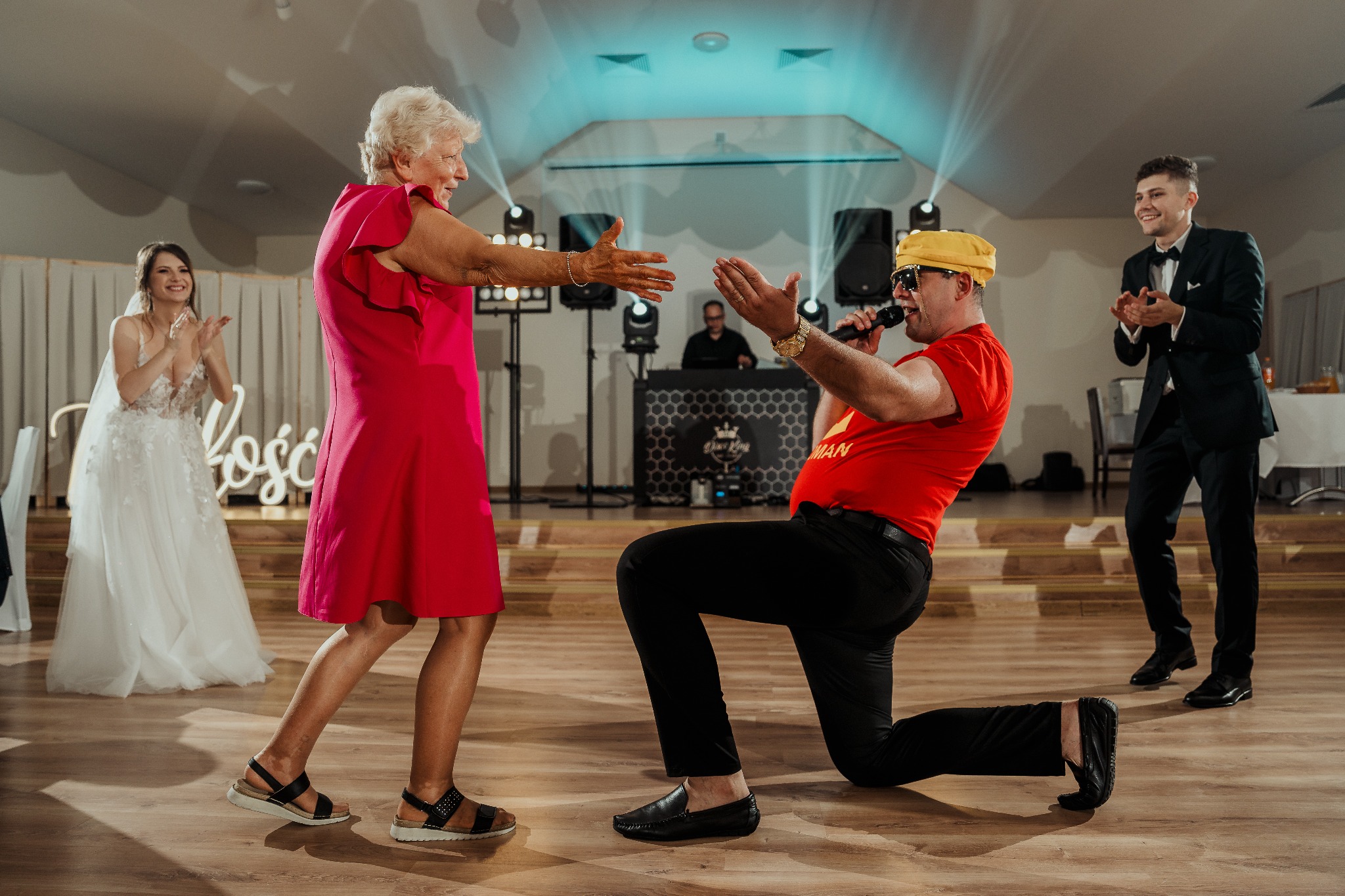 Występ zespołu na weselu: wokalista w żółtym turbanie klęczy przed gościem, w tle DJ, nagłośnienie i oświetlenie. Para młoda i goście klaszczą.