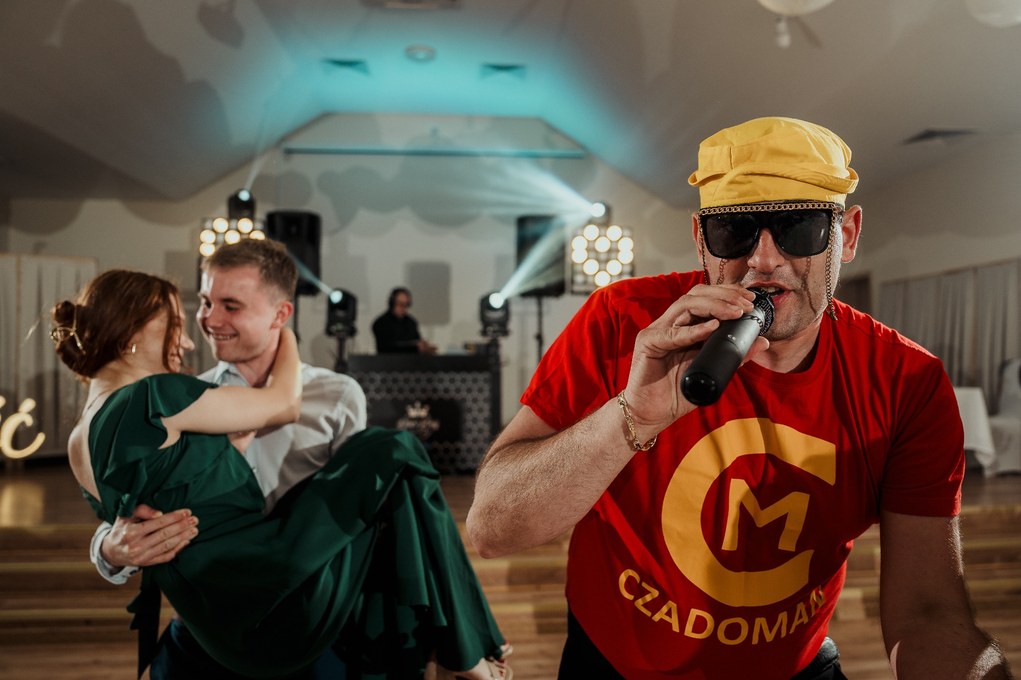 Wodzirej w czerwonej koszulce z mikrofonem na tle bawiących się gości weselnych i oświetlenia scenicznego. Zabawa integracyjna z klasą i humorem.