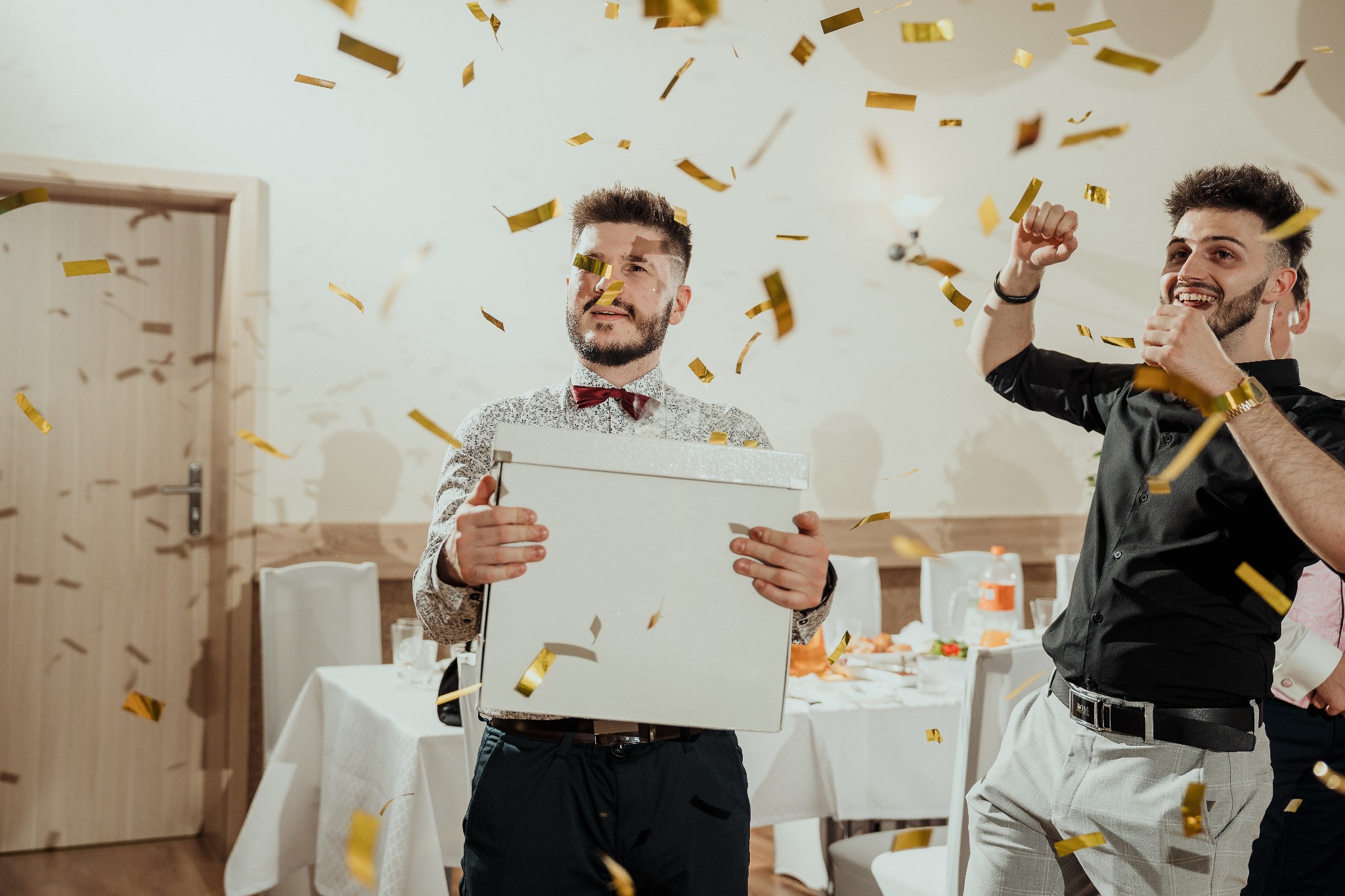 Dwóch mężczyzn w trakcie oczepin na weselu, złote konfetti w powietrzu. Jeden trzyma białe pudełko, drugi w euforii, tło: białe stoły i krzesła.