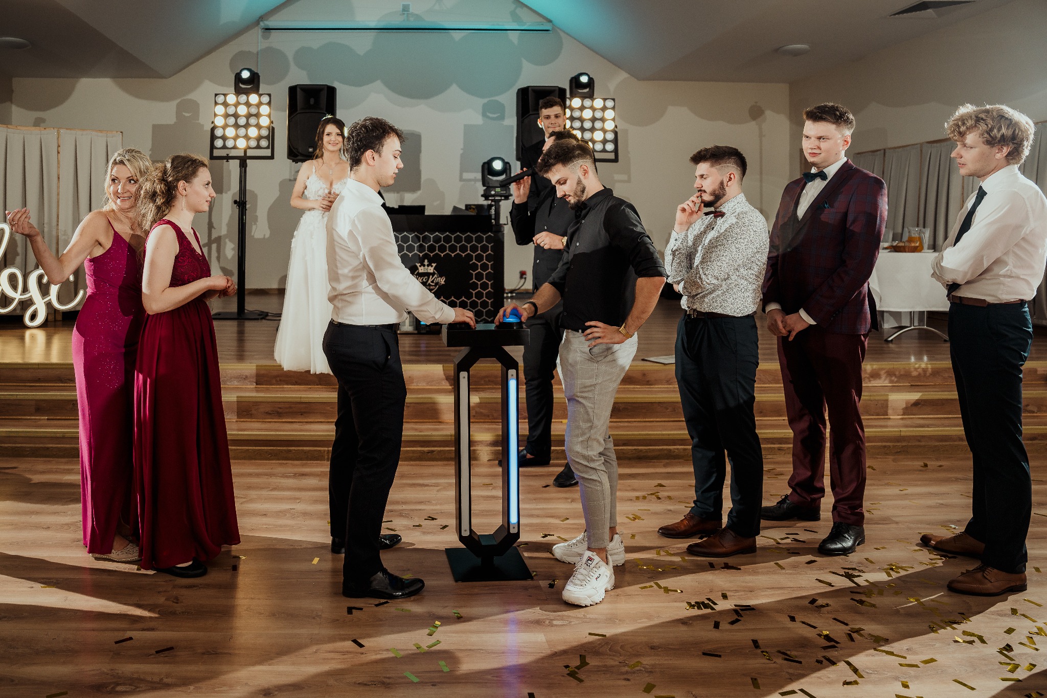 Goście weselni w eleganckich strojach otaczają interaktywny panel DJ'a z podświetleniem, na tle sceny z oświetleniem i parą młodą. Parkiet pokryty konfetti.