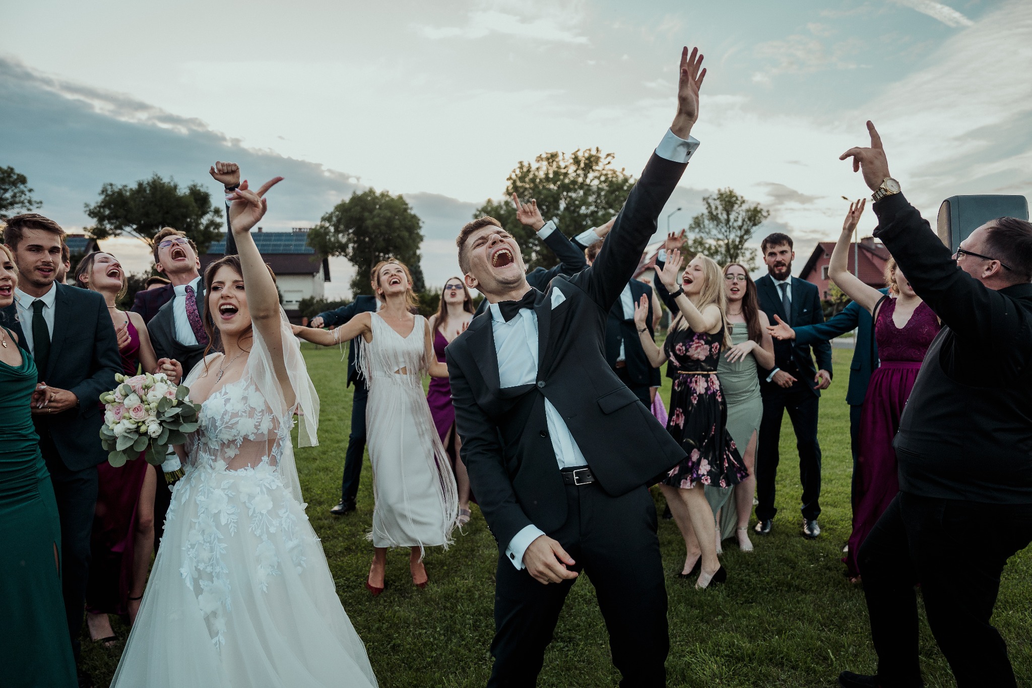 Rozbawieni goście weselni tańczą na trawie, w tle niebo. Panna młoda z bukietem, pan młody w muszce, wszyscy z uniesionymi rękami w radosnym geście.