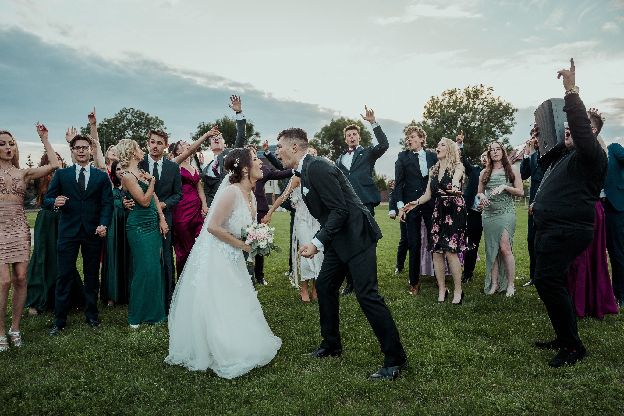 Rozbawieni nowożeńcy z gośćmi na zielonej trawie, jeden z gości trzyma głośnik. Dynamiczne ujęcie z pleneru podczas imprezy weselnej.