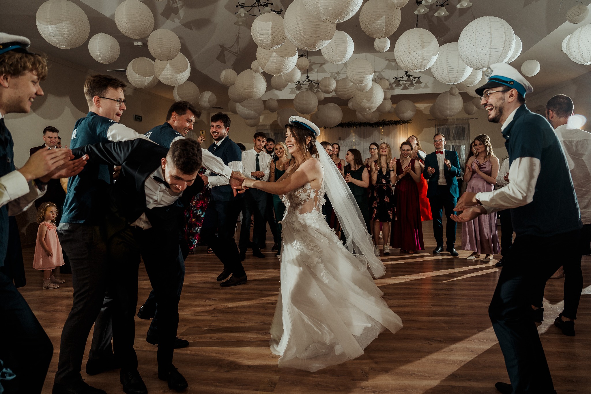 Radosna zabawa weselna: panna młoda w tańcu prowadzona przez gości w marynarskich czapkach, tło wypełnione papierowymi lampionami. Parkiet i uśmiechnięci goście.