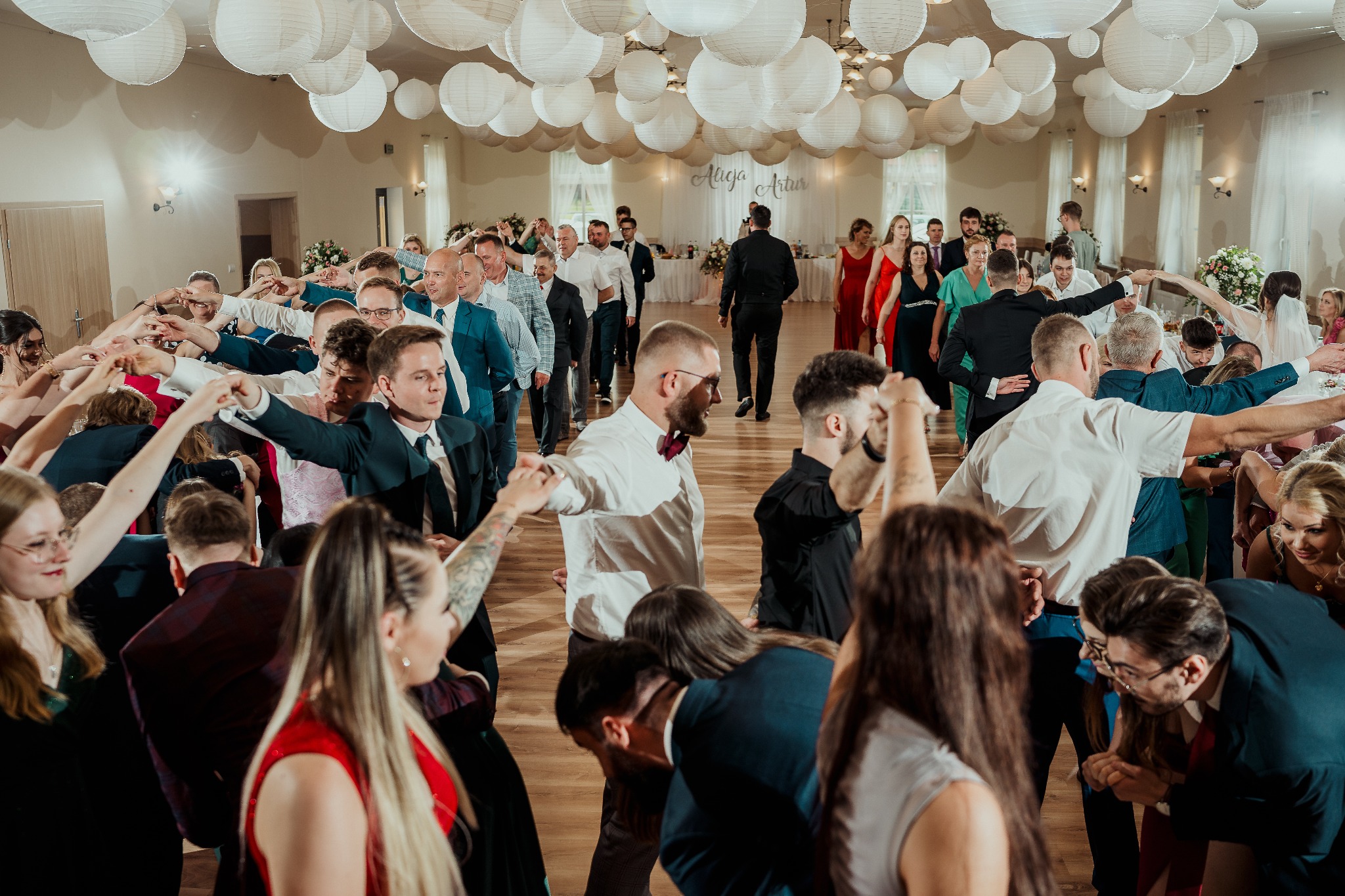 Goście weselni tworzą tunel dla pary młodej w sali z dekoracjami z białych lampionów. Widoczni ludzie w różnym wieku i ubraniach, tworzący szpaler.