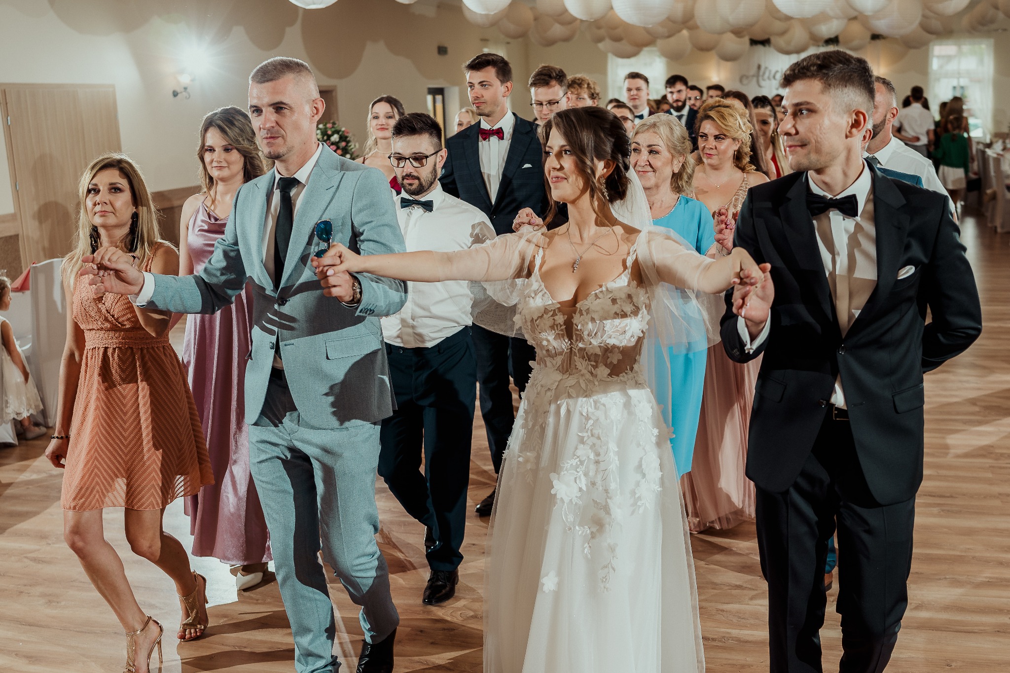 Korowód weselny na parkiecie: para młoda w centrum, goście w eleganckich strojach, rustykalny klimat sali z dekoracjami i drewnianą podłogą, uśmiechnięte twarze.