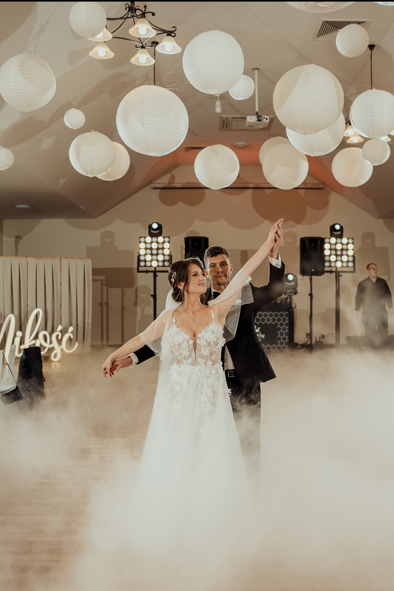 Para młoda tańczy pierwszy taniec w dymie, oświetlona światłami i lampionami. W tle widoczne nagłośnienie i napis 'Miłość' z neonu.