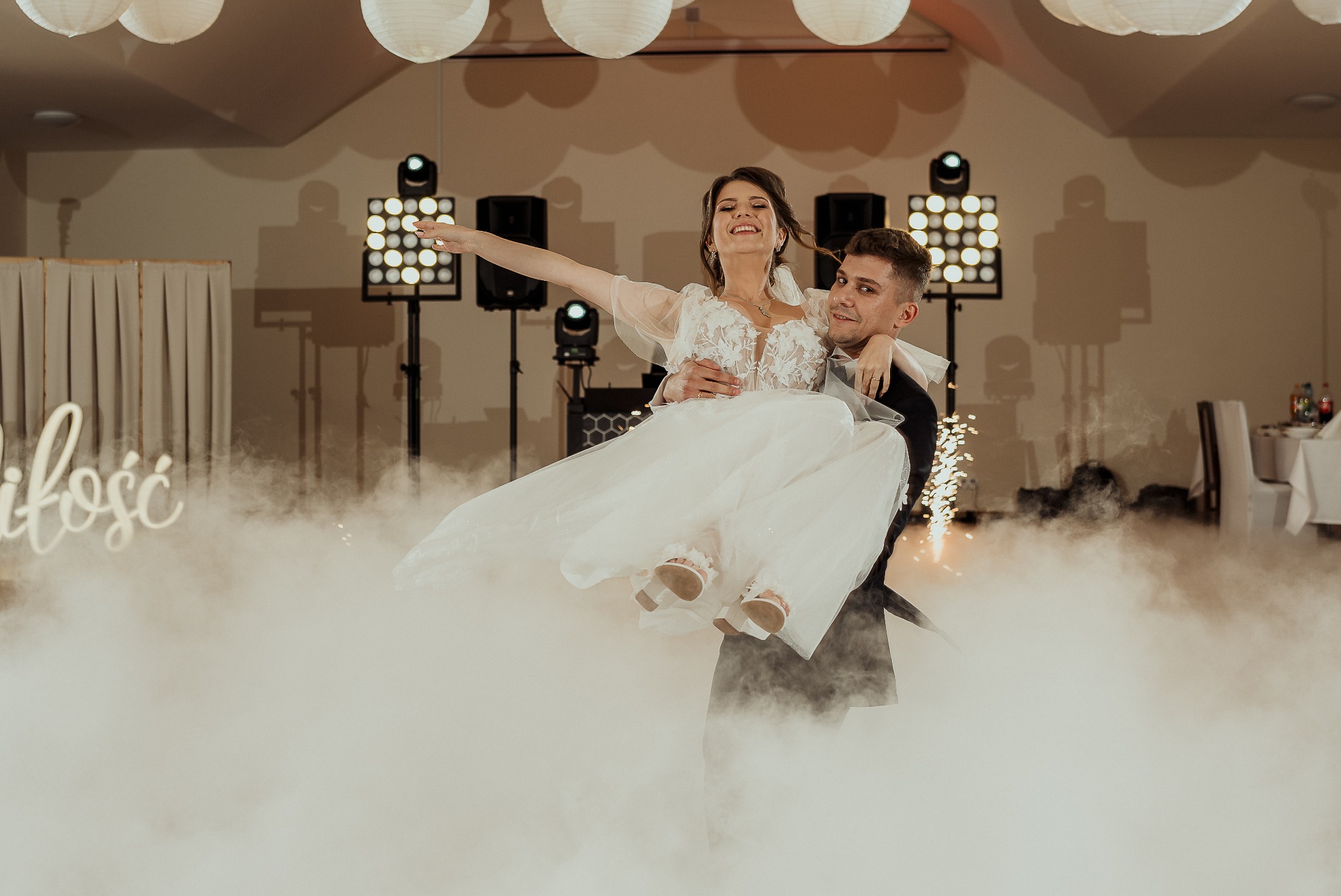 Para młoda na parkiecie podczas wesela, unoszona przez pana młodego w objęciach, w tle oświetlenie sceniczne i dekoracje, efekt mgły na podłodze.