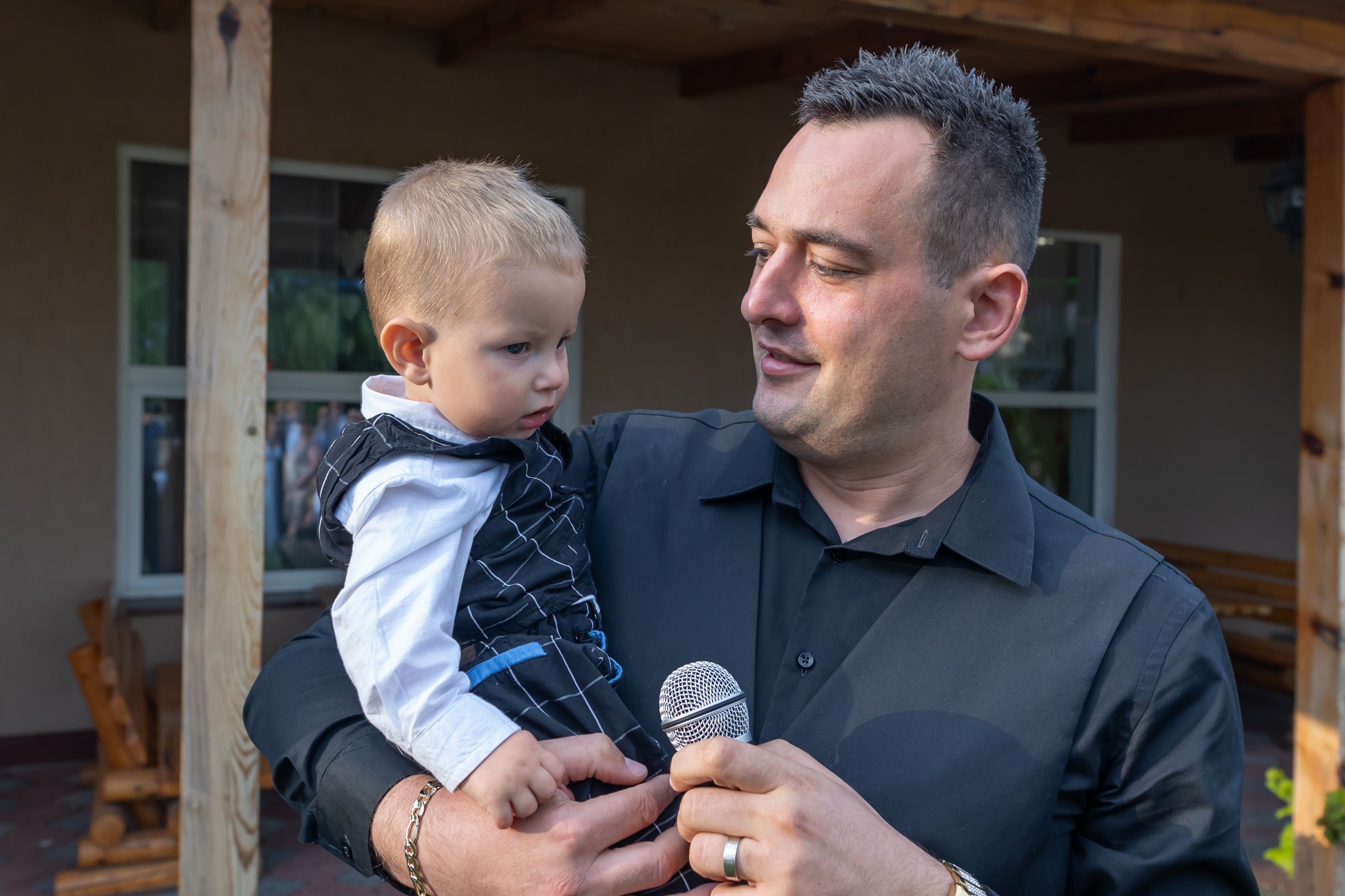 Prezenter z mikrofonem trzyma na rękach małego chłopca w eleganckiej kamizelce. Uśmiech na twarzy mężczyzny, w tle budynek z oknami.