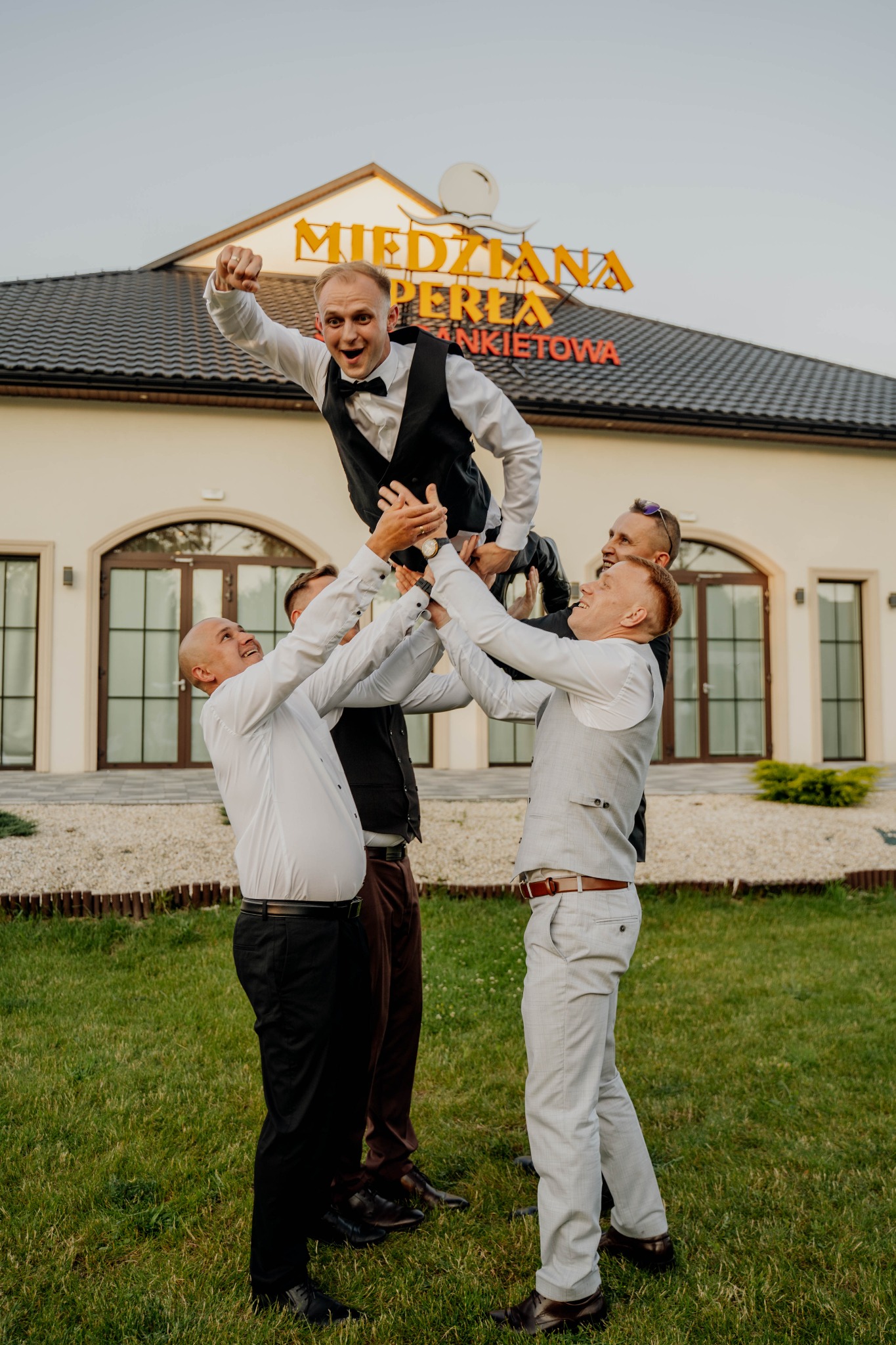 Ekipa weselna podrzuca uśmiechniętego pana młodego na tle budynku z napisem 'Miedziana Perła'. Wszyscy w eleganckich strojach, wesoła atmosfera.