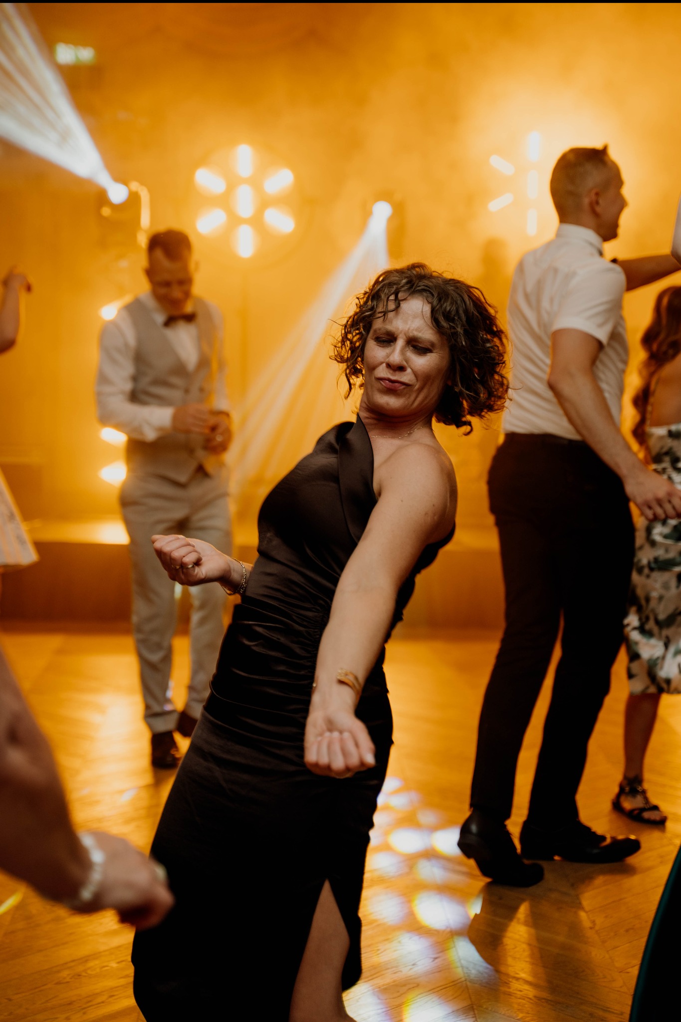 Roztańczona kobieta w czarnej sukni na parkiecie, w tle zespół muzyczny i goście weselni, oświetlenie sceniczne w ciepłych barwach.