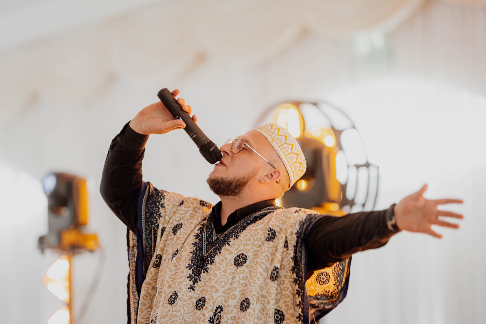 Wokalista z brodą, w ozdobnym stroju i czapce, śpiewa do mikrofonu, rozkładając ręce na tle scenicznego oświetlenia.