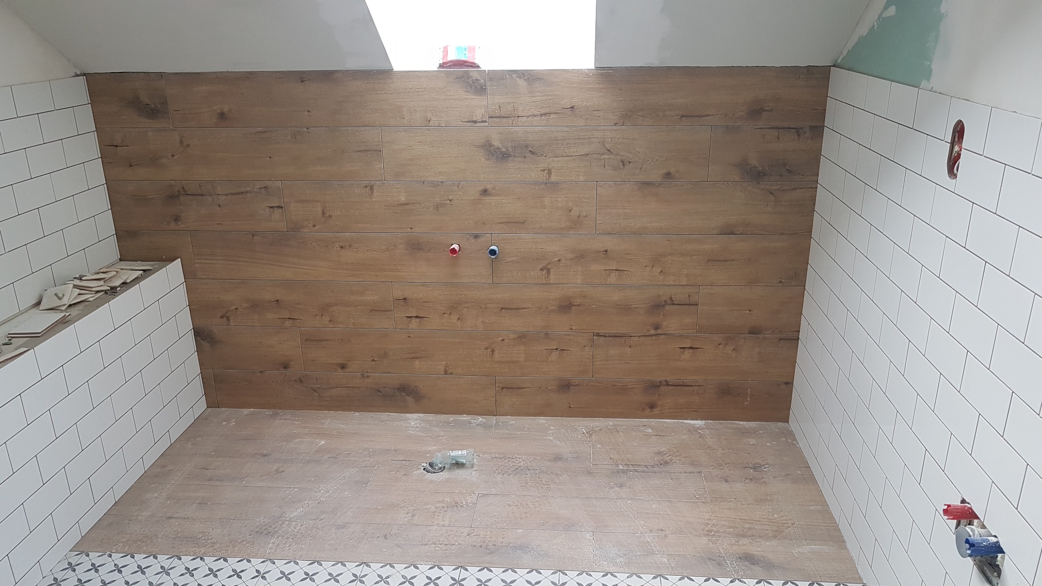 Łazienka w trakcie wykańczania: ściana z płytek imitujących drewno, białe płytki na pozostałych ścianach, widoczne otwory na baterie, fragment podłogi z wzorzystym wykończeniem.