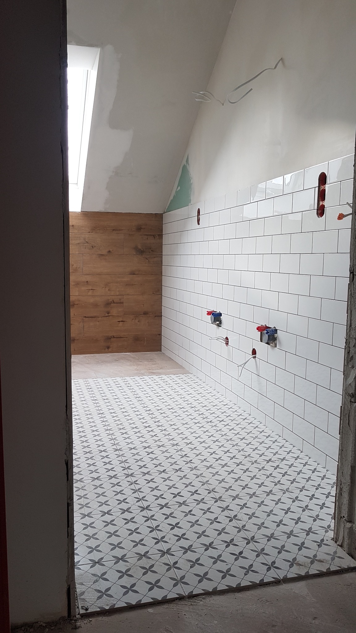 Wnętrze łazienki w trakcie remontu: białe, prostokątne płytki na ścianie z widocznymi instalacjami wodnymi, podłoga wyłożona wzorzystą, szaro-białą ceramiką, drewnopodobne wykończenie jednej ze...