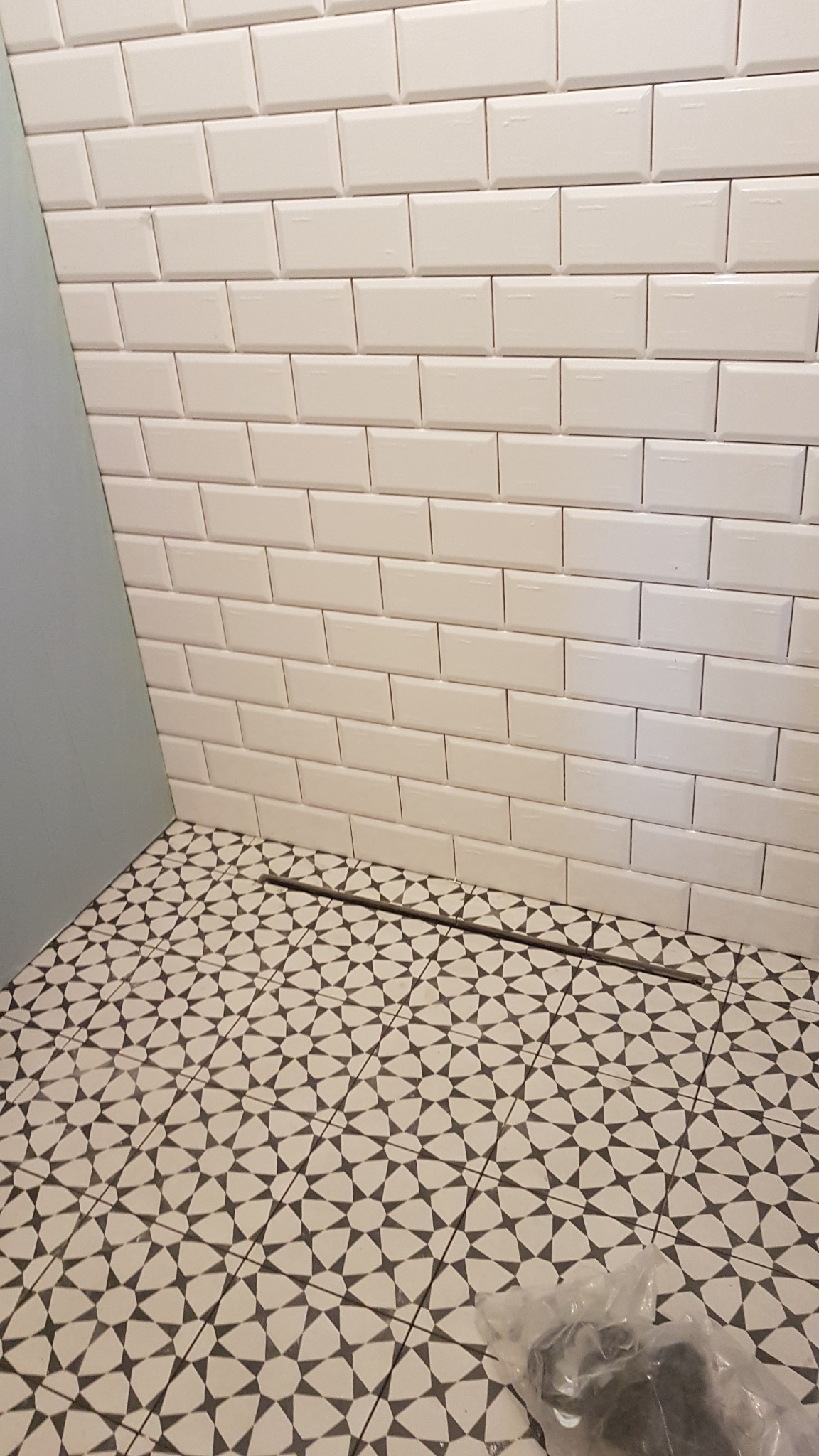 Białe, prostokątne płytki ścienne ułożone w cegiełkę nad wzorzystą, biało-czarną podłogą w łazience, widoczny odpływ liniowy i fragment niebieskiej ściany.