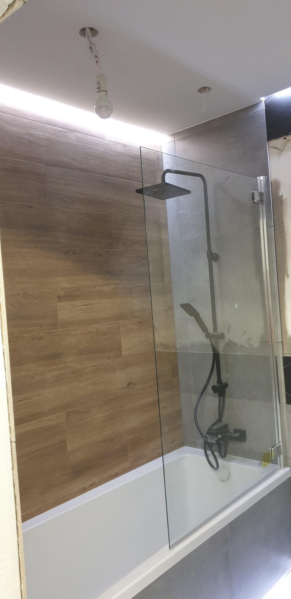 Nowoczesna kabina prysznicowa z czarną armaturą i szklanym panelem zamontowana nad białą wanną, płytki imitujące drewno na ścianie, surowa żarówka zwisająca z sufitu.