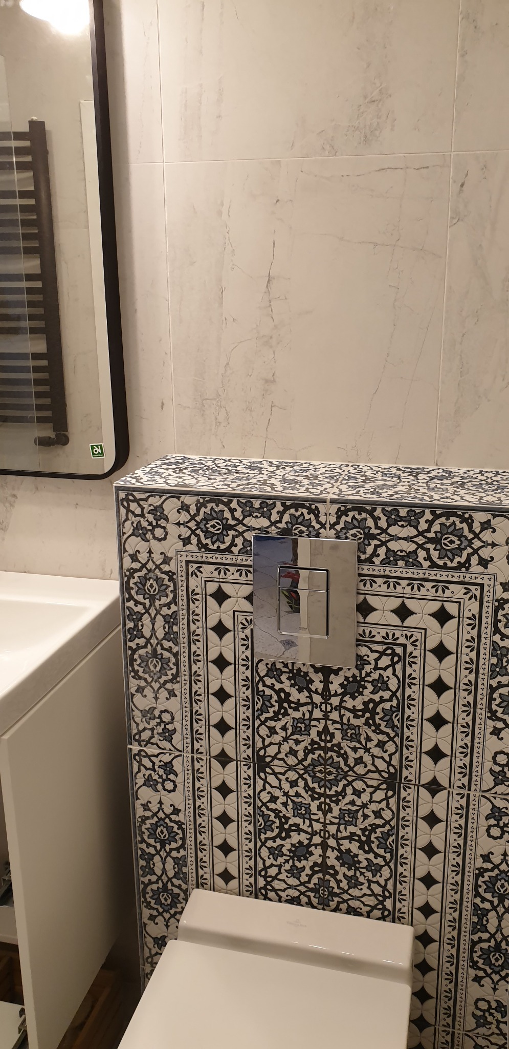 Kompakt WC z zabudową w płytkach dekoracyjnych z motywem kwiatowym, chromowany przycisk spłukiwania, biała umywalka i grzejnik drabinkowy w tle.