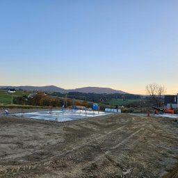 Roboty ziemne Jaskulski Jarosław - Wylana płyta fundamentowa z wystającymi prętami zbrojeniowymi, widoczna ubikacja TOI TOI, mini wywrotka i teren przygotowany pod budowę na tle pagórkowatego krajobrazu o zmierzchu.