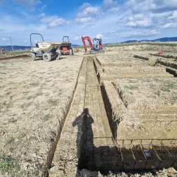 Roboty ziemne Jaskulski Jarosław - Przygotowanie terenu pod fundamenty: wykopane rowy z widocznym zbrojeniem, na drugim planie maszyny budowlane - wywrotka i mini koparka, błękitne niebo z chmurami w tle.